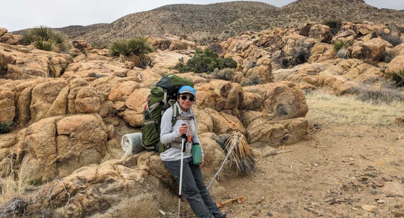 A person wearing a backpack and carrying a trekking pole leans against a rock in a desert environment. 